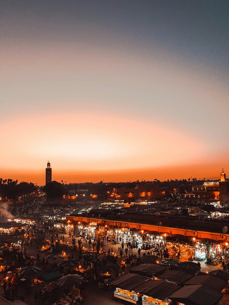 Cette photo surélevée de Jemaa el-Fnaa capture l'énergie vibrante de la place de Marrakech pendant un « premier voyage au Maroc », un lieu de rencontre historique et culturel incontournable. 
Les stands du marché, éclairés par des lampes chaudes et des rangées de auvents sombres. 
