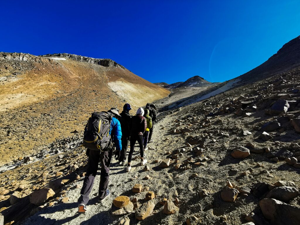 Des randonneurs gravissent un sentier de haute montagne rocheux et aride sous un ciel bleu profond, en direction du village berbère d'Imlil niché au fond de la vallée.