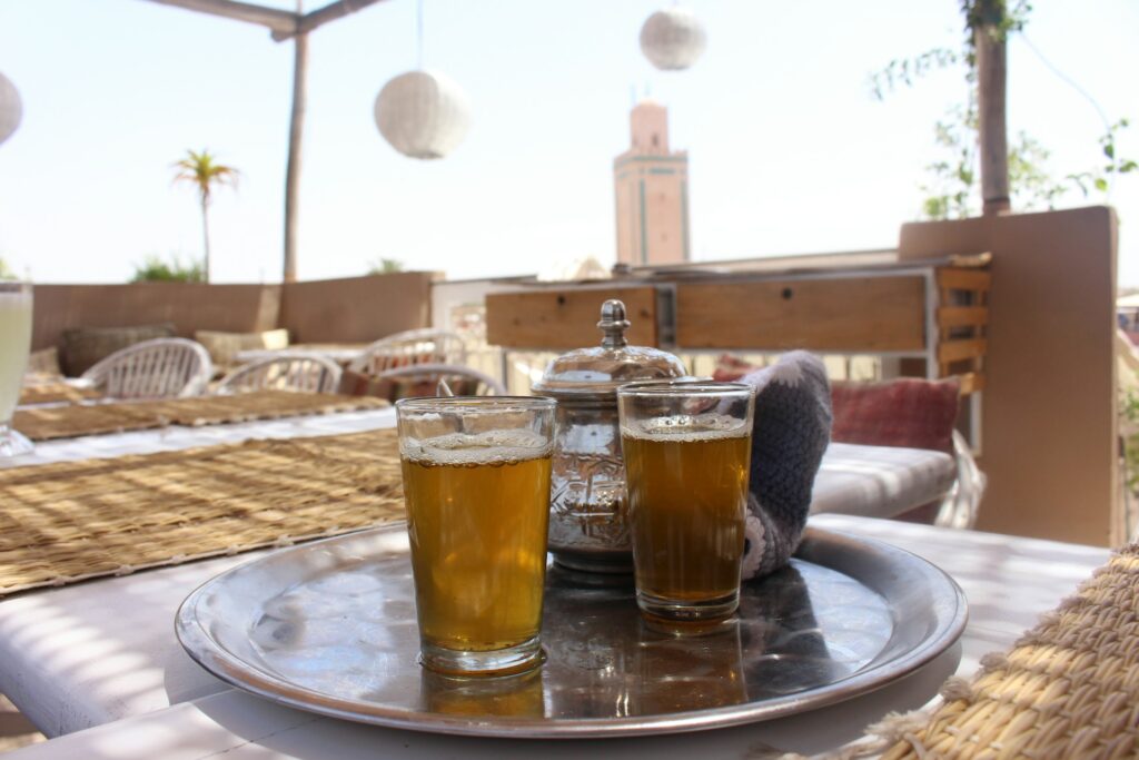 Verres de thé à la menthe servis sur un plateau traditionnel sur une terrasse ensoleillée au Maroc, avec vue sur un minaret et une ambiance typique marocaine.