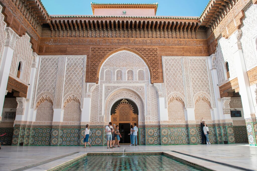 Cour intérieure richement décorée d’un monument historique au Maroc, avec mosaïques zellige, sculptures traditionnelles et visiteurs admirant l’architecture marocaine.