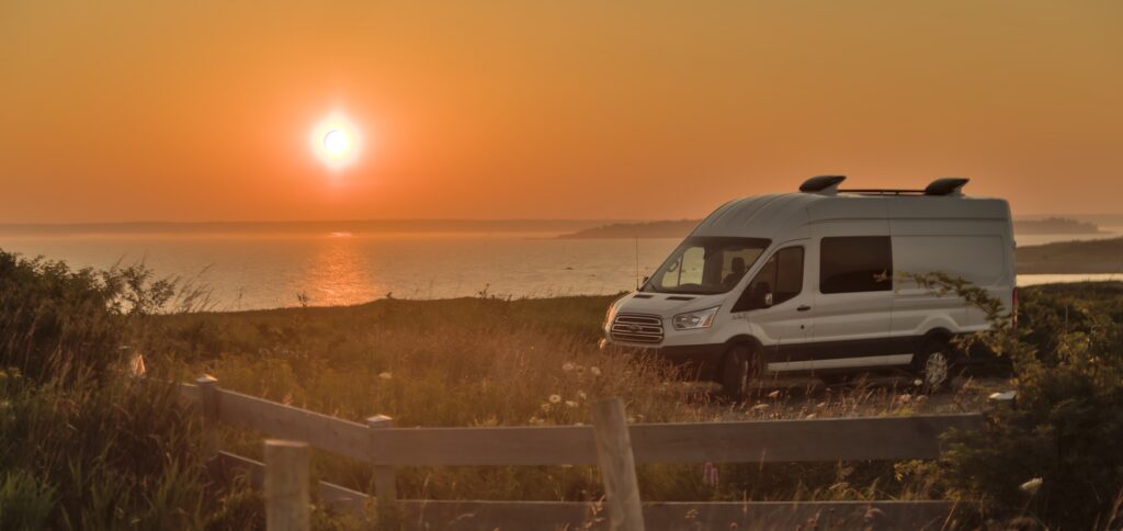 Un camping-car stationné face à un magnifique coucher de soleil, installé dans un paysage naturel où la lumière dorée se reflète sur l’eau. Cette scène évoque la liberté du voyage nomade et l’importance de préparer son camping-car aux micro-climats Marocains avant de partir à l’aventure. 