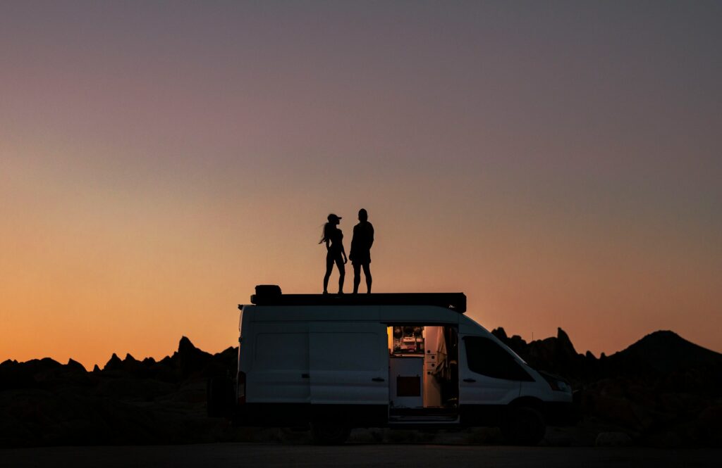 L'image montre un camping-car ou un fourgon aménagé. En silhouette sur le toit du véhicule se tiennent deux personnes, un homme et une femme, observant le paysage.  Préparer son camping-car aux micro-climats Marocains est une étape essentielle pour garantir un voyage agréable et sans encombre