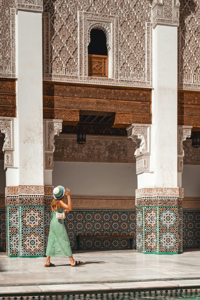  Une femme, vue de dos et vêtue d'une robe longue vert pastel mouchetée et d'un chapeau de soleil à rayures bleues et blanches, marche sur le sol en marbre blanc d'une cour intérieure de style mauresque. Cela représente comment préparer sa valise pour partir au Maroc en 2026
