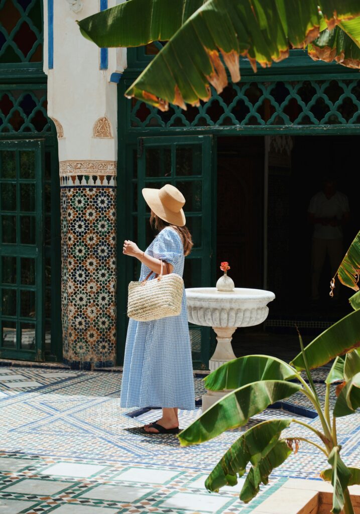  Une femme, vue de dos et vêtue d'une robe longue vert pastel mouchetée et d'un chapeau de soleil à rayures bleues et blanches, marche sur le sol en marbre blanc d'une cour intérieure de style mauresque. Cela représente comment préparer sa valise pour partir au Maroc en 2026