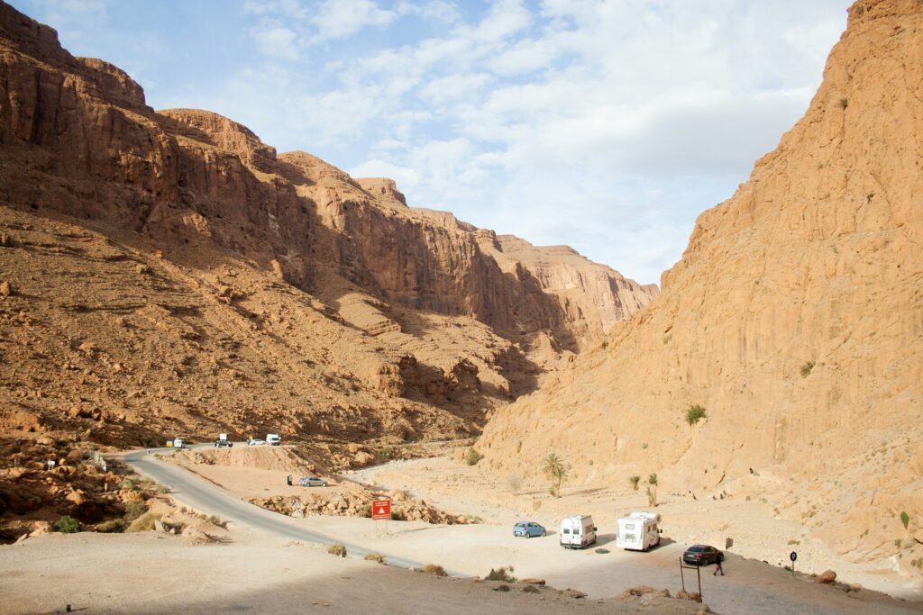 Une image panoramique et légèrement élevée capture la gorge de Todra au Maroc, bordée d'imposantes falaises de grès brun. Une route pavée sinueuse s'étend dans la vallée. L'image illustre une façon populaire de visiter le pays, en profitant de l'indépendance qu'offre le fait de parcourir le Maroc en camping-car.
