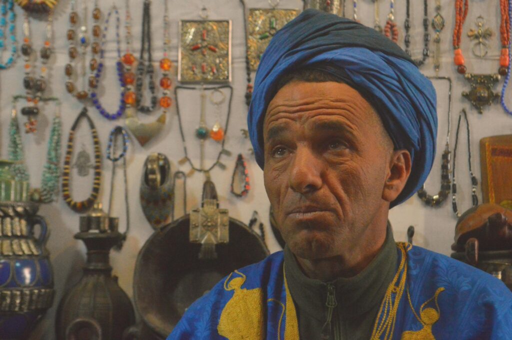Portrait d’un artisan marocain dans un souk traditionnel, avec bijoux et objets artisanaux en arrière-plan, illustrant les rencontres camping-car Maroc au cœur de l’artisanat local.