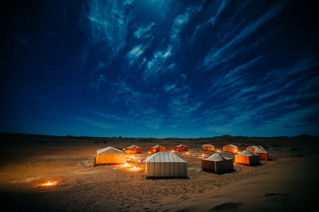 Sous un ciel nocturne d’un bleu profond parsemé d’étoiles et de nuages filamenteux, un campement de tentes traditionnelles s’illumine doucement. Cette atmosphère magique et sereine évoque parfaitement les Excursions dans le Sahara marocain, où l’on découvre la beauté brute du désert sous les étoiles.