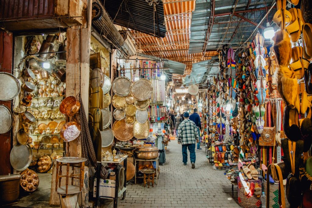Cette image illustre une ruelle animée des souks de la médina de Marrakech, où les échoppes débordent d'objets artisanaux marocains. Cette scène met parfaitement en valeur l'importance de suivre quelques Conseils pratiques avant de visiter Marrakech.