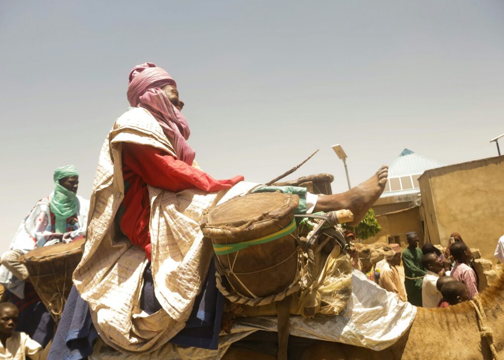 Sur cette photographie prise lors du festival international des nomades, un homme vêtu de vêtements traditionnels colorés (turban rose, tunique rouge et grand drapé beige) est assis sur un chameau. Il tient un grand tambour en peau et lève fièrement un pied nu, souriant. 