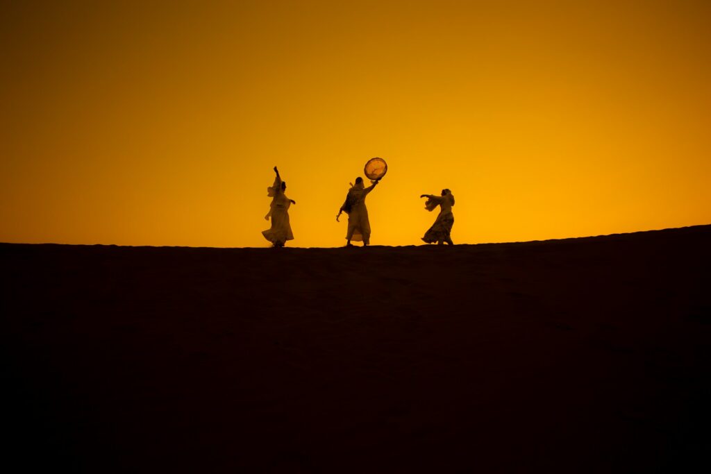 Trois silhouettes dansent sur la crête d’une dune, se découpant nettement sur un ciel orange flamboyant au coucher du soleil. Cette scène poétique et vibrante a été capturée lors du festival international des nomades, célébrant la culture et les traditions des peuples du désert.