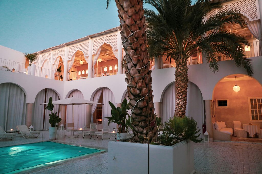 Découvrez les Riads au Maroc via ce magnifique patio blanc aux palmiers, piscine turquoise scintillante et lumières chaleureuses du crépuscule.